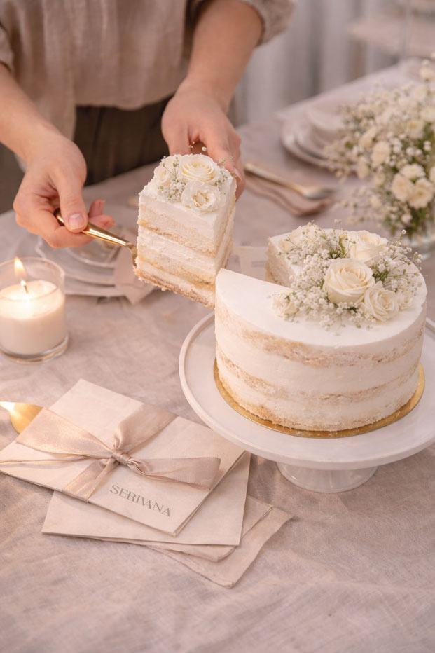 A person using a gold server to lift a slice of tiered white cake decorated with cream roses and baby's breath.