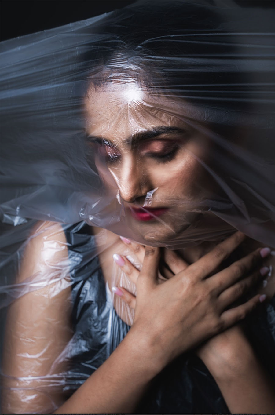 A dramatic, conceptual portrait of a woman with her face and shoulders partially obscured by a layer of thin, crinkled plastic film