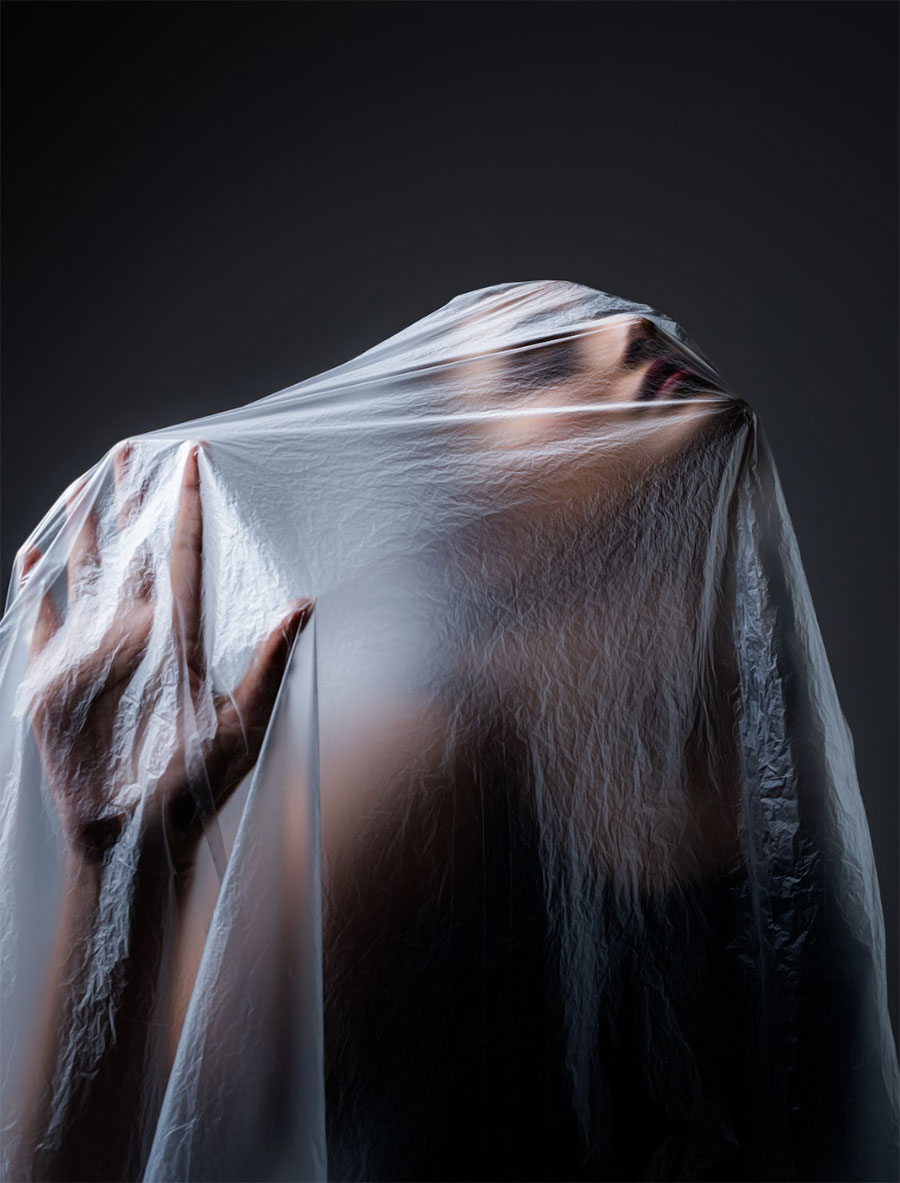 A dramatic, conceptual portrait of a woman with her face and shoulders partially obscured by a layer of thin, crinkled plastic film