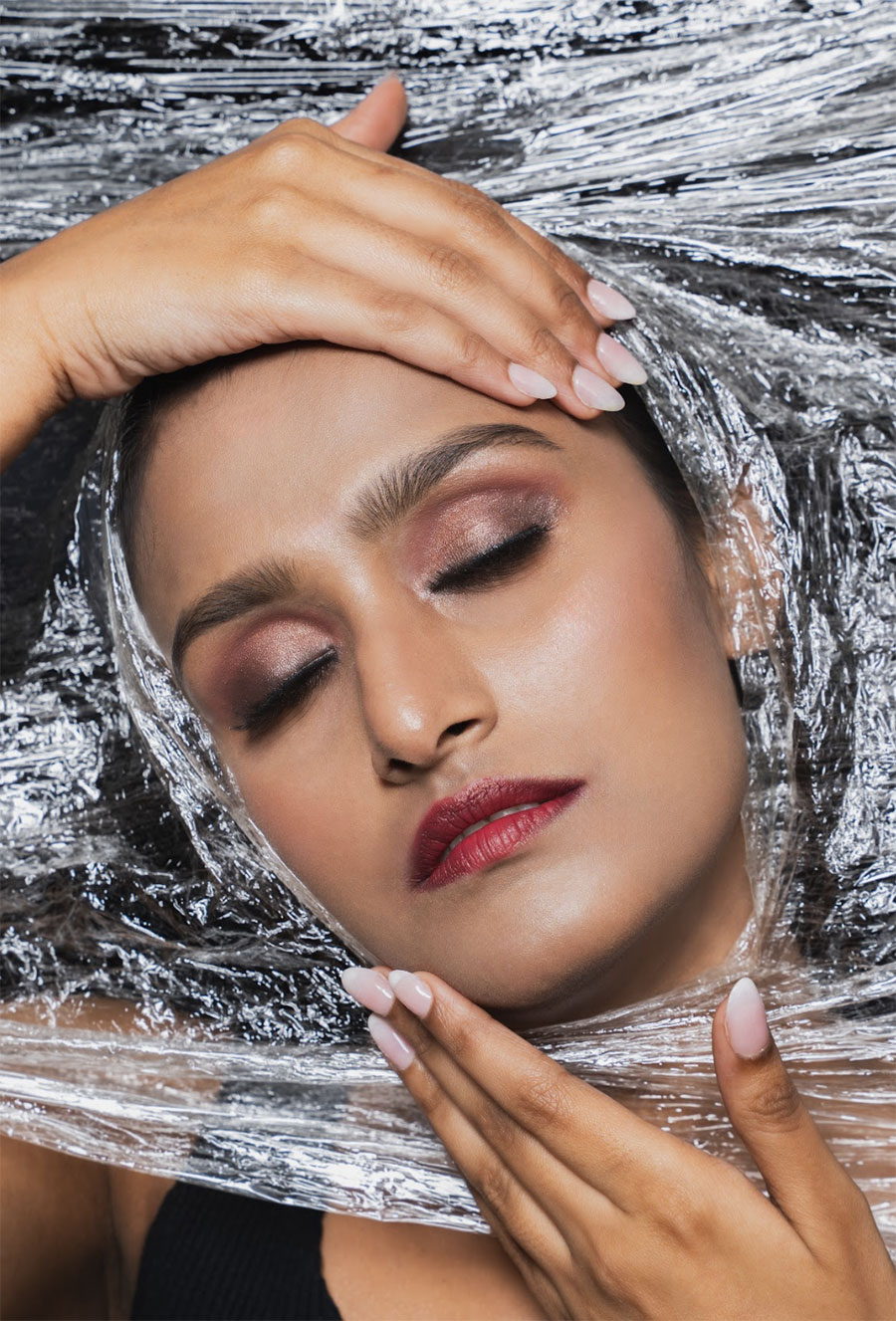 An artistic, close-up fashion portrait of a person with eyes closed, framed by the textured, crinkled surface of a reflective plastic film that envelops the subject’s head and hands